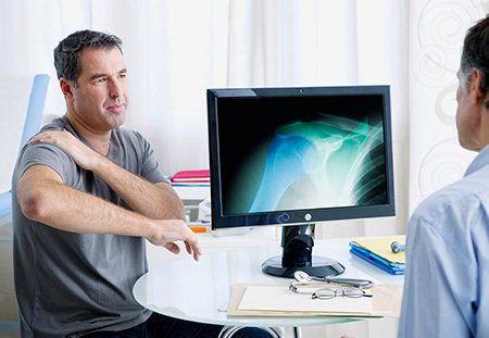 A man undoubtedly telling his doctor his shoulder hurts while the doctor looks at an xray of the fractured shoulder bone.